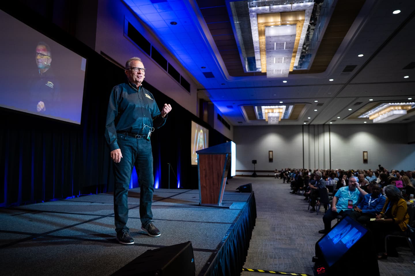 Mike Wilk, President and Founder of Five Rings Financial, speaking on stage to a large audience at an FRF event.